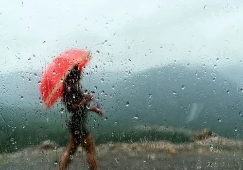 A woman with a red umbrella walking outside as seen on a window with droplets of rain