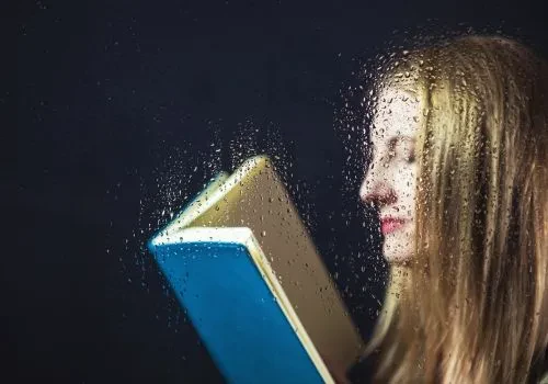 A woman reading a book by the rainy window sill